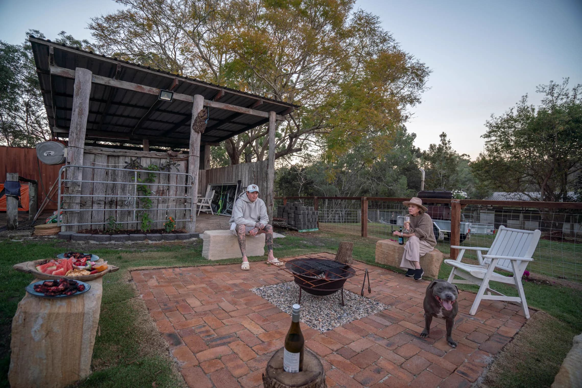 Two guests and a dog around the lit fire pit at dusk, a platter and wine on the log tables.