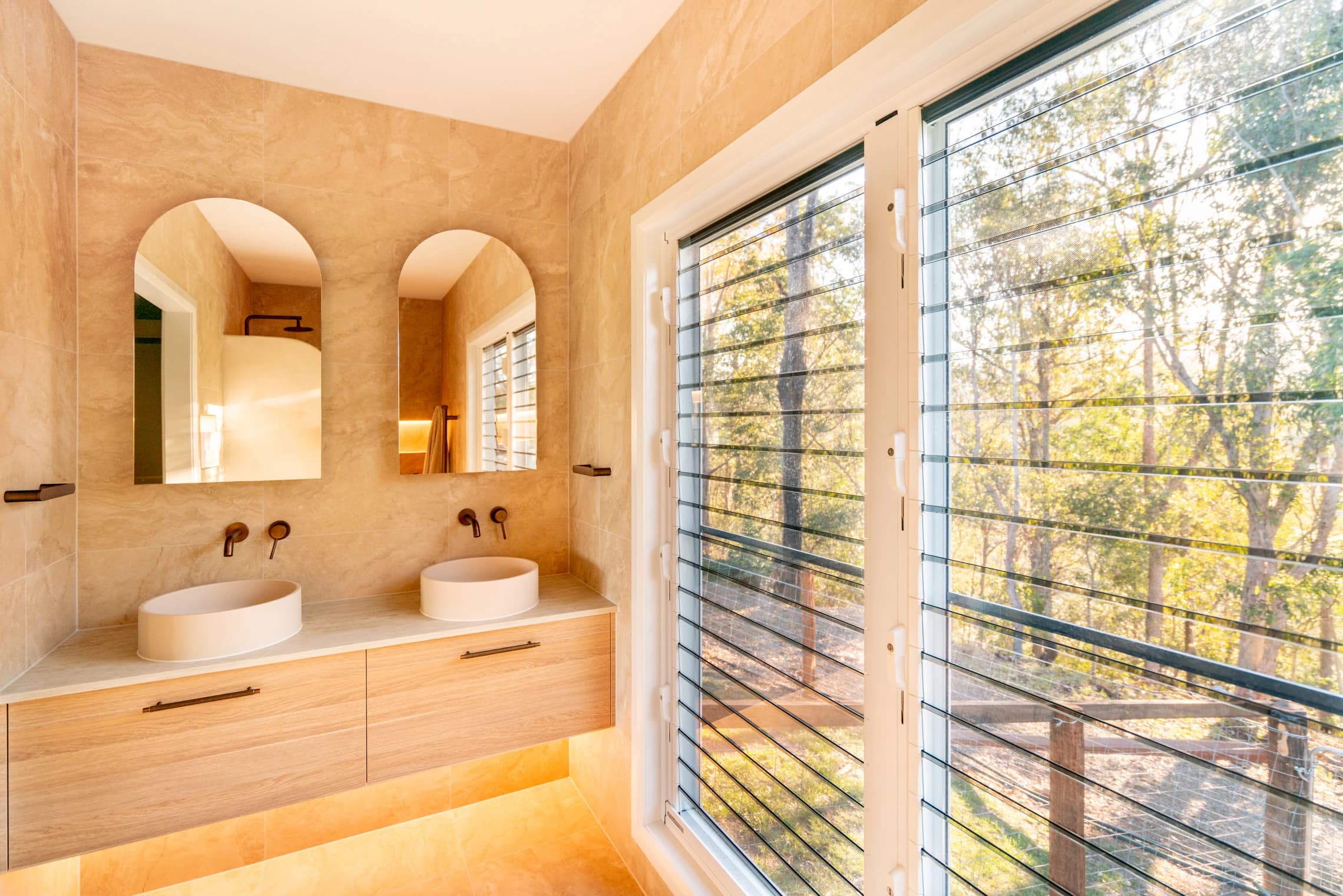Twin basins and arched mirrors beside a louvre window overlooking trees.