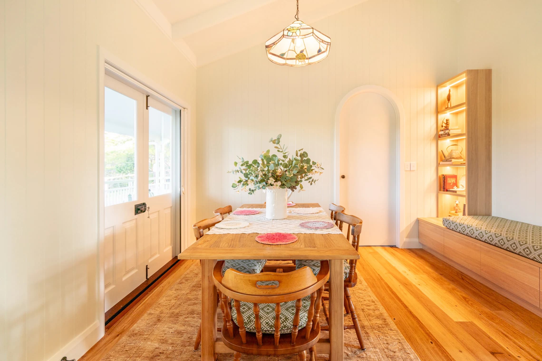 A timber dining table beneath a stained-glass pendant beside the window bench.